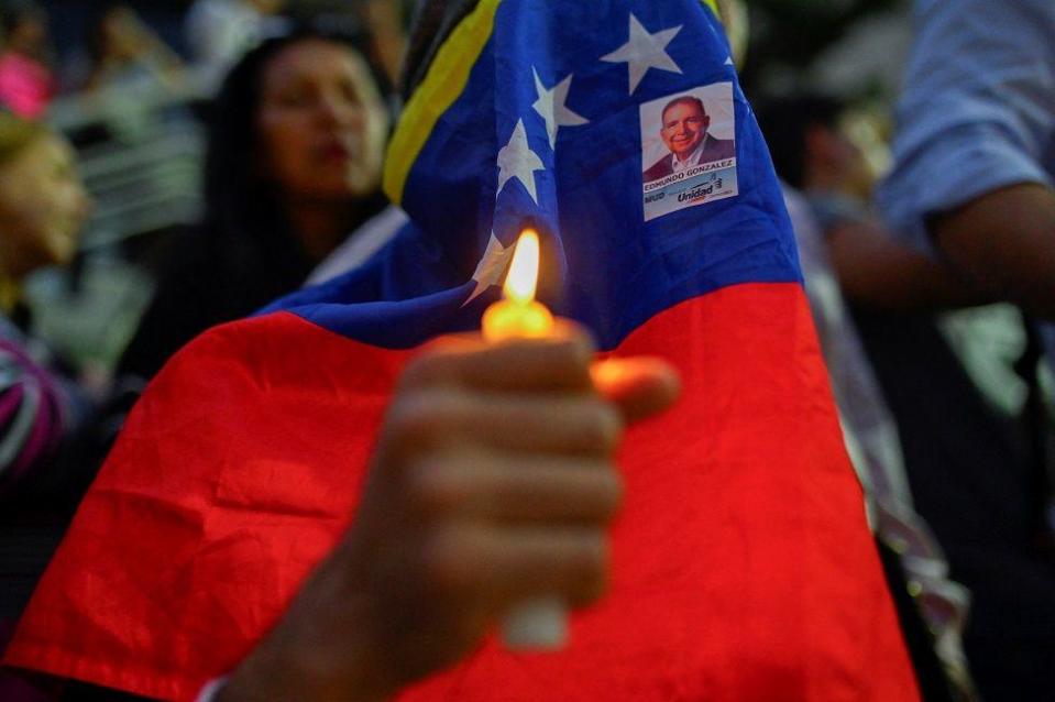 Uma pessoa com uma vela acesa e o rosto coberto pela bandeira da Venezuela e pela foto de Edmundo González