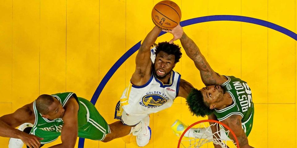 An overhead shot shows Andrew Wiggins leaping in the air and grabbing a rebound while surrounded by Celtics players.