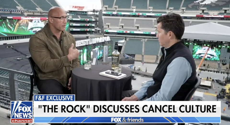 Dwayne "The Rock" Johnson in a conversation with a reporter in a stadium setting, discussing cancel culture
