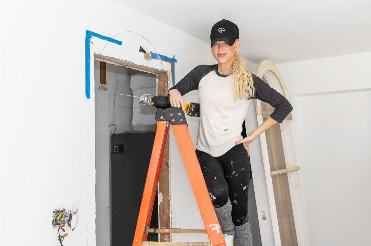 galey alix of hgtv's home in a heartbeat standing on ladder wearing paint spattered t shirt jeans ball cap