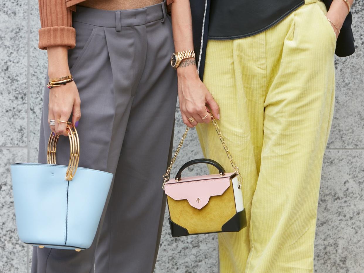 two women standing next to each other on the street wearing trousers and carrying small purses