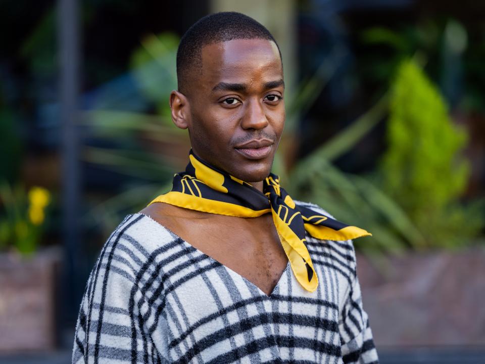 Actor Ncuti Gatwa wears checkered black white v neck top, black pants, purple bag, yellow black scarf outside JW Anderson during London Fashion Week February 2023 on February 19, 2023 in London, England