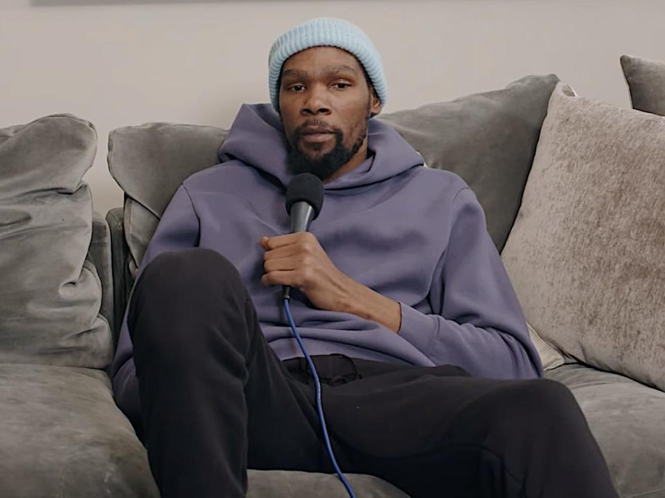 Kevin Durant sits on a couch and holds a microphone while speaking on a podcast.