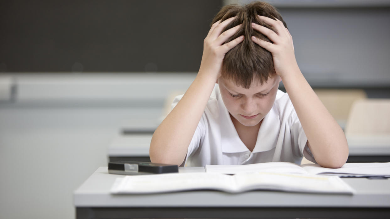 A student, seemingly frustrated with his hands placed on top of his head, looks down at his schoolwork.
