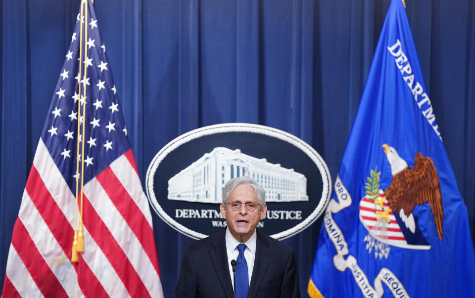 Merrick Garland at a microphone in front of a blue curtain, flags and a Justice Department seal.