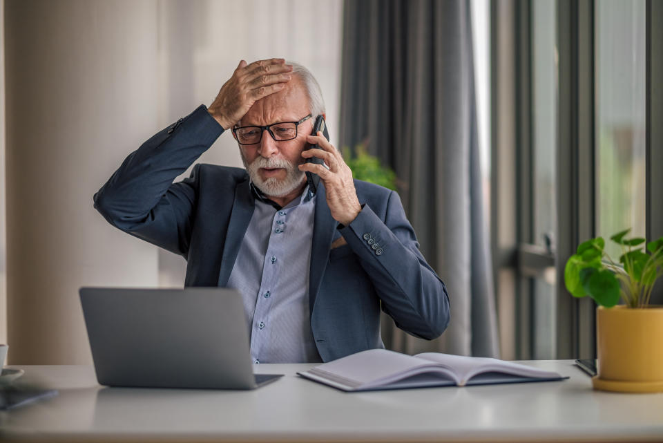 Anxious businessman talking on mobile phone while working on laptop. Worried senior male professional is wearing formals. He is sitting at desk in corporate office.