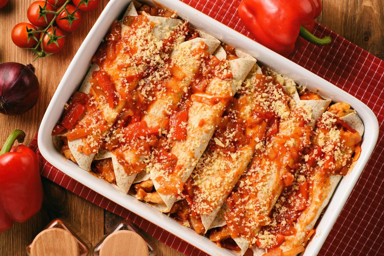 Top view of six chicken enchiladas in a ceramic rectangle white pan on a red table mat on a wooden table, surrounded by red bell peppers, cherry tomatoes, and an onion