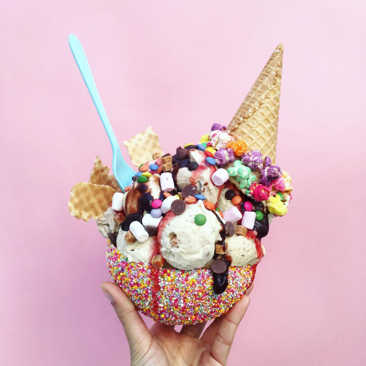 Female hand holding an edible ice cream bowl with tons of different ice cream and toppings on a light pink-purple background