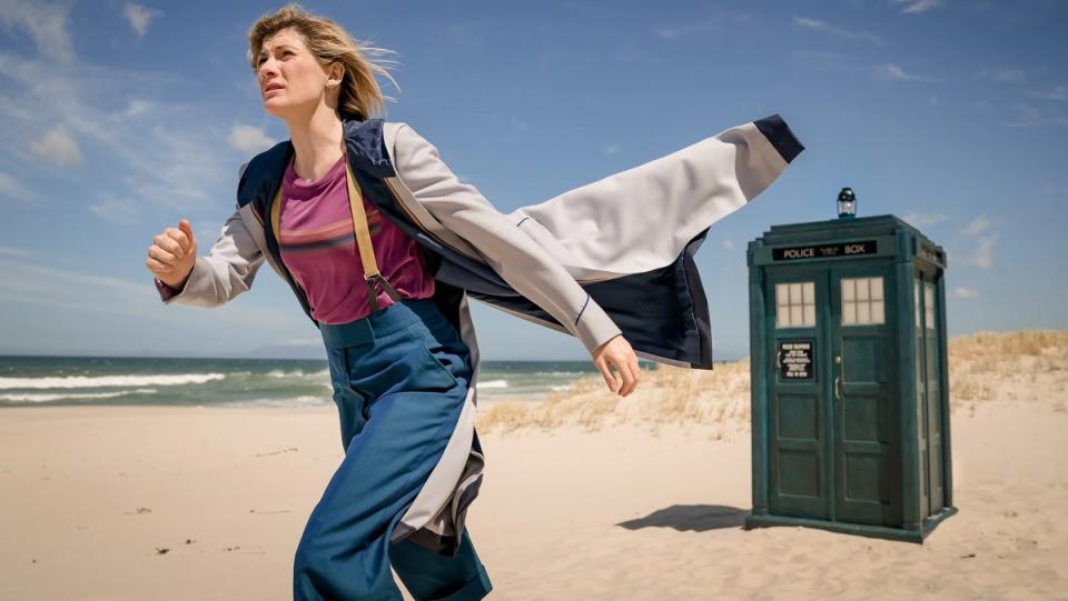 Jodie Whittaker in her usual Thirteenth Doctor costume of high-waisted trousers, suspenders, a t-shirt, and a long raincoat, walks away from the TARDIS on a sunny beach near a body of water.