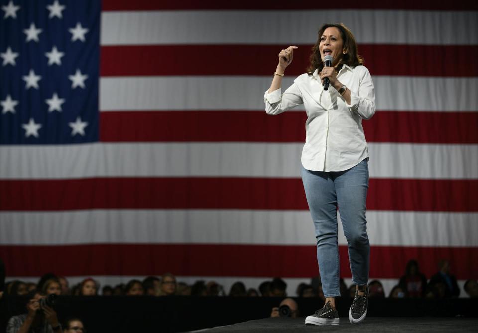 Woman wearing white shirt, jean and sneakers speaks on a microphone flanked by an American flag.