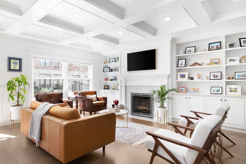 ELMHURST, IL, USA - MARCH 30, 2021: A luxurious, white living room with a coffered ceiling, a television mounted above a tiled fireplace, and built in shelving in front of chairs and a couch