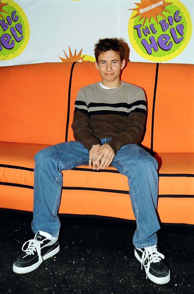 JTT in a sweater, jeans, and sneakers and smiling on the couch