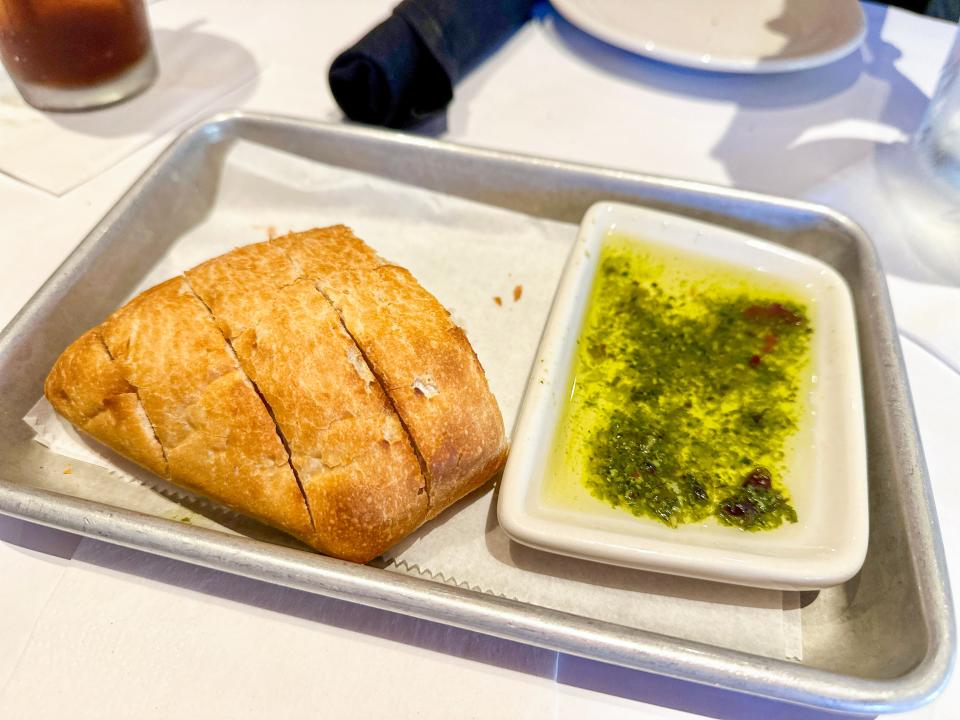 A piece of crusty bread with four slices cut out and an olive-oil dipping sauce with herbs in a dish next to it. The food sits on a silver serving tray