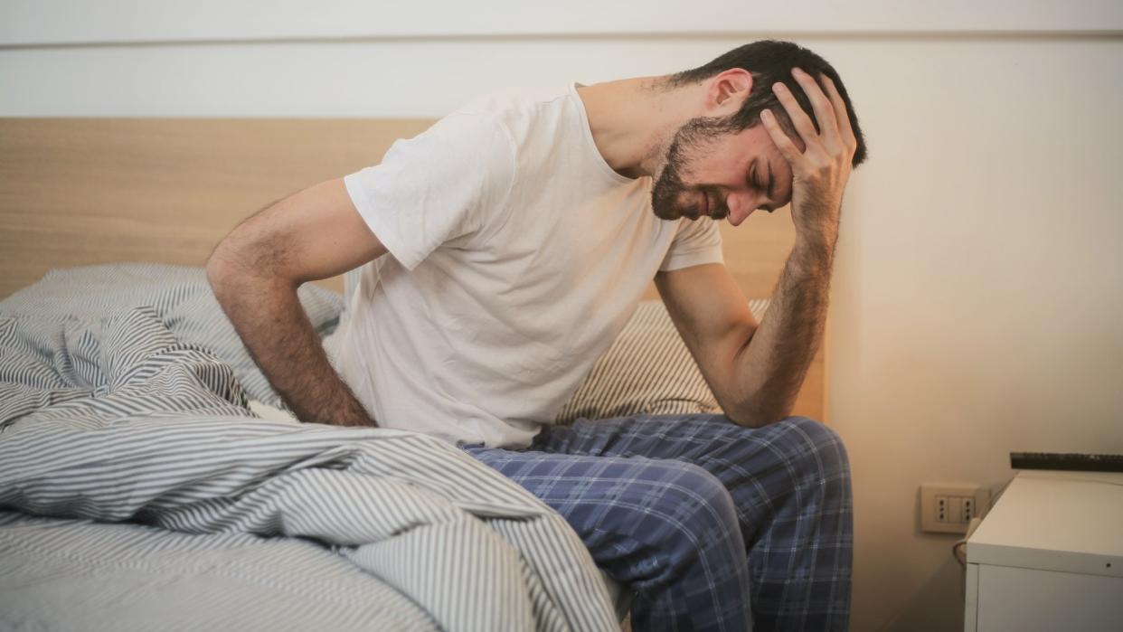  A man sits on his bed holding his head and back because back pain woke him up 