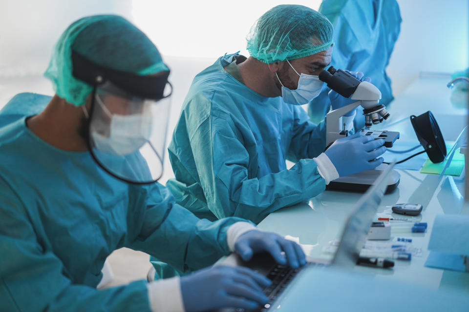 Medical workers in hazmat suit working with laptop computer and micoroscope inside laboratory hospital during coronavirus outbreak - Focus on center man face