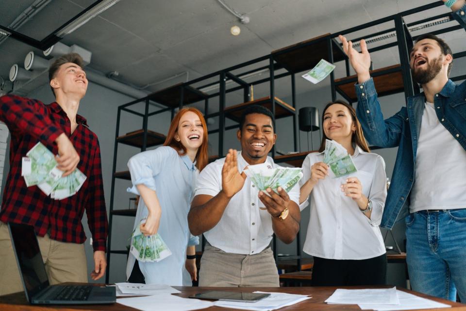 A group of five people smiling with fistfuls of cash.