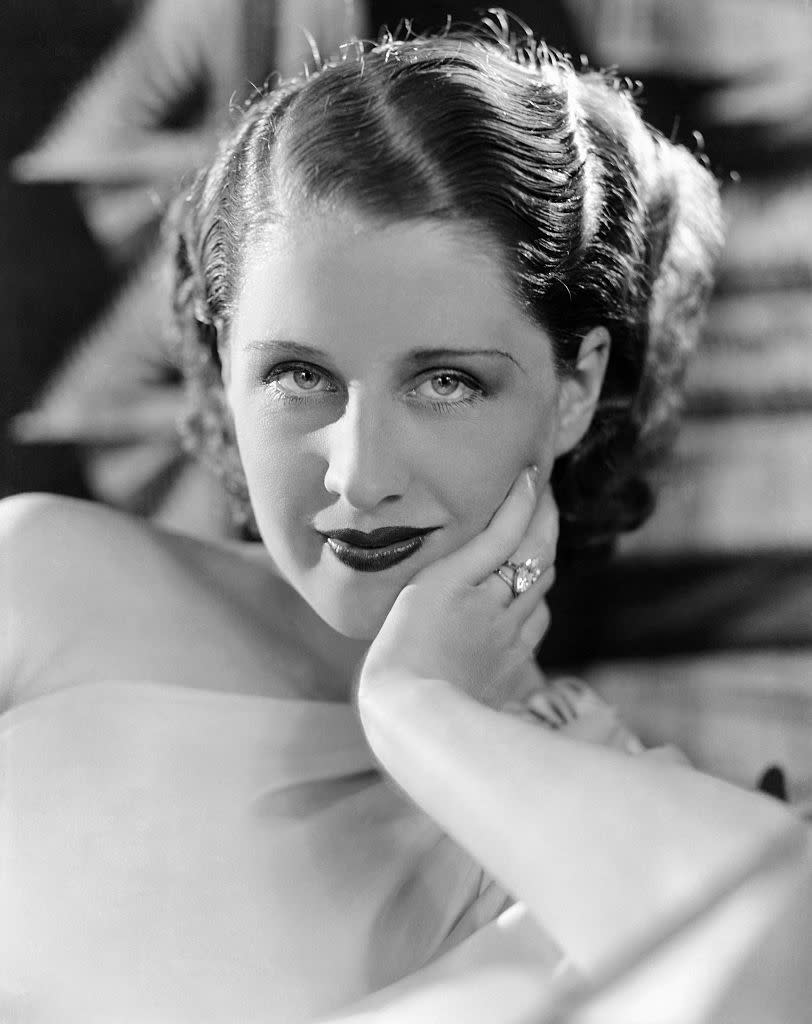 A close-up portrait of a woman with wavy, short hair, resting her chin on her hand, wearing a ring and light-colored clothing