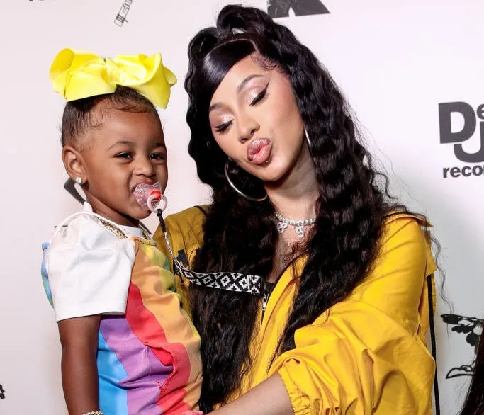 Cardi B in a draped outfit holding her daughter, who's in a colorful dress with a yellow bow in her hair