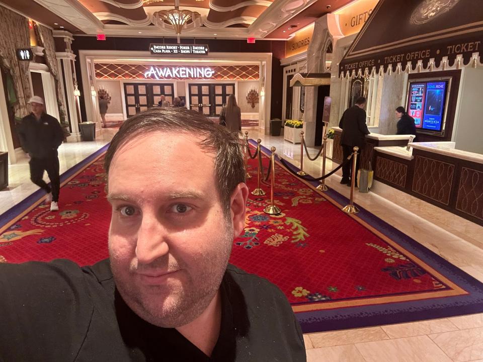 A man taking a selfie in front of a theater box office area for a show called "Awakening."