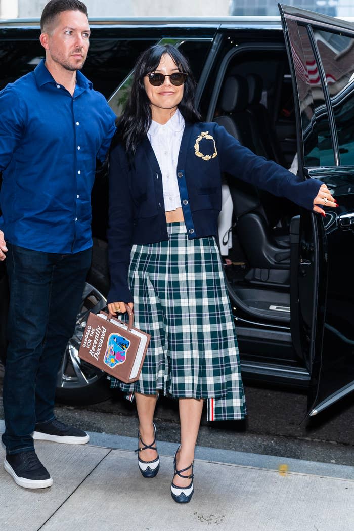 Jenna Ortega steps out of a car holding a book-shaped clutch, wearing a plaid skirt, white blouse, and a navy cardigan. An unidentified man stands nearby