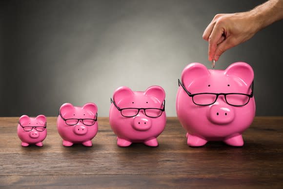 A row of progressively larger pink piggy banks wearing glasses with a man's hand holding a coin over the largest.