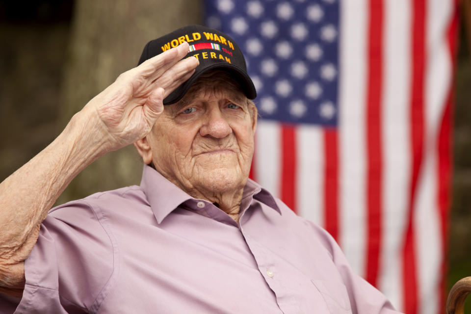 An elderly World War II veteran salutes, wearing a black cap with "World War II Veteran" written on it. An American flag is in the background