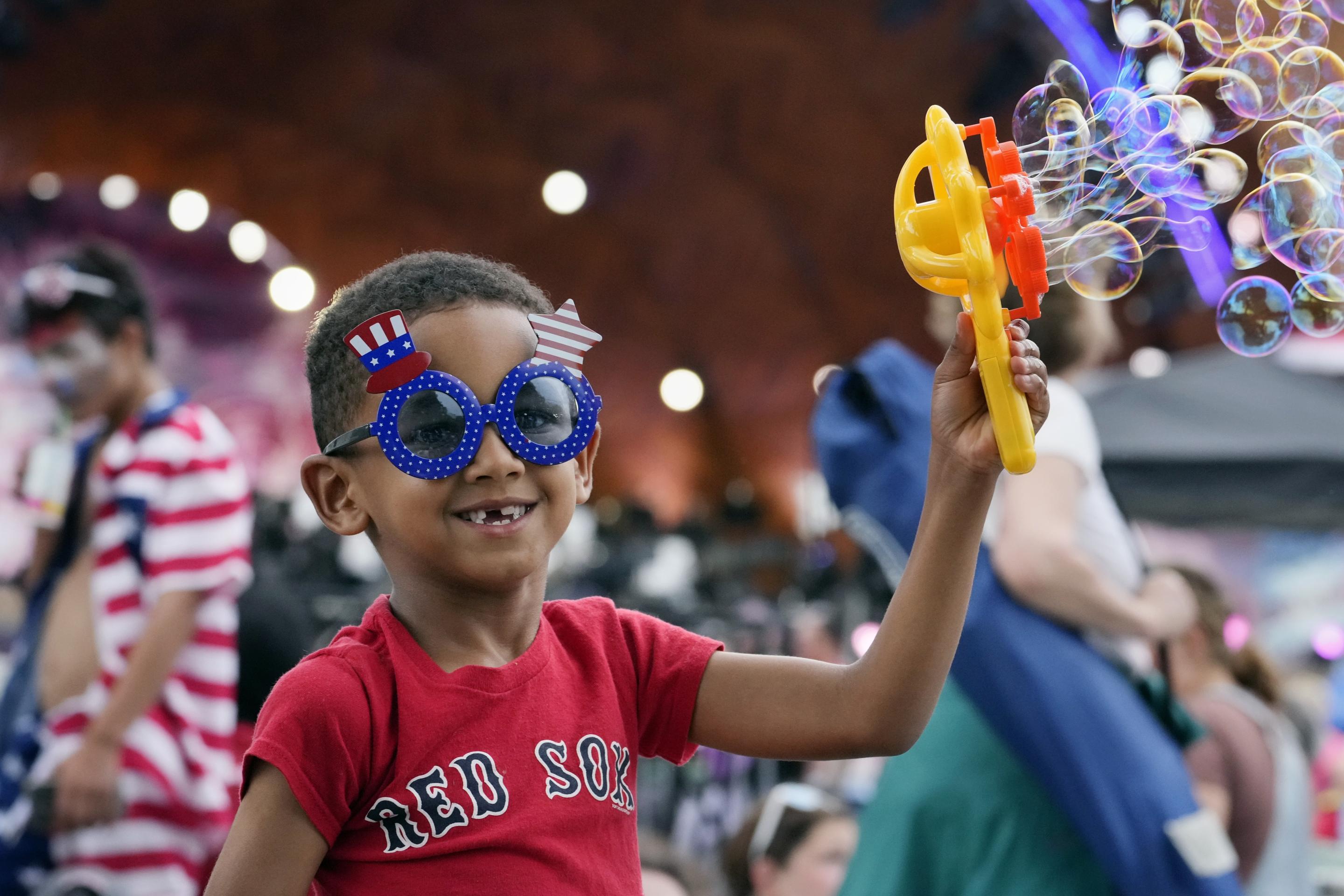 A young boy wearing a Red Sox shirt and Fourth of July-theme sunglasses holds a bubble machine.