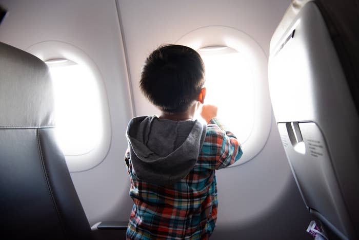 A young child in a plaid shirt looks out from a plane window, holding onto the frame