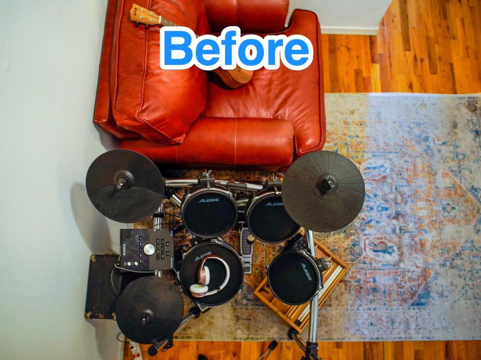 Blue text says "before" at the top of an aerial photo of a small square room with wood floors, a grey rug, a red leather love seat, and a black electric drum kit.