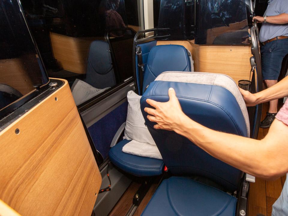 Two staggered bus seats with a pillow on one of the seats as someone adjusts the other seat.