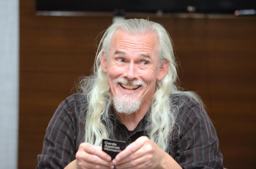 A man with long silver hair in a dark long-sleeve shirt holding a black card in his hands and smiling