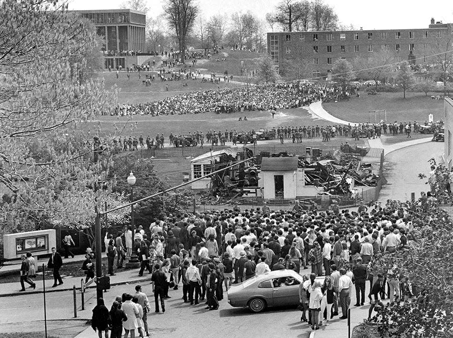 A crowd of students, some carrying their books gathers near the charred remains of the ROTC building on the Kent State campus. At the top left is Taylor Hall, where, around noon on the building’s far side, National Guardsmen clashed with protesters and opened fire.