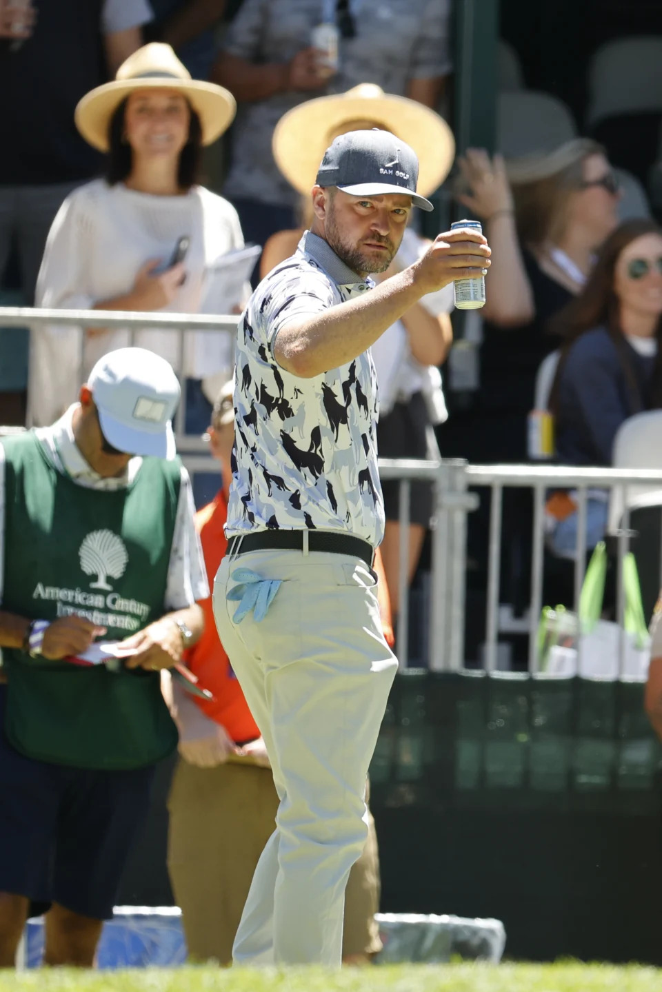 A man holding a drink at an outdoor event dressed in a patterned golf shirt, white pants, and a cap, with a crowd in the background, including one person wearing a hat