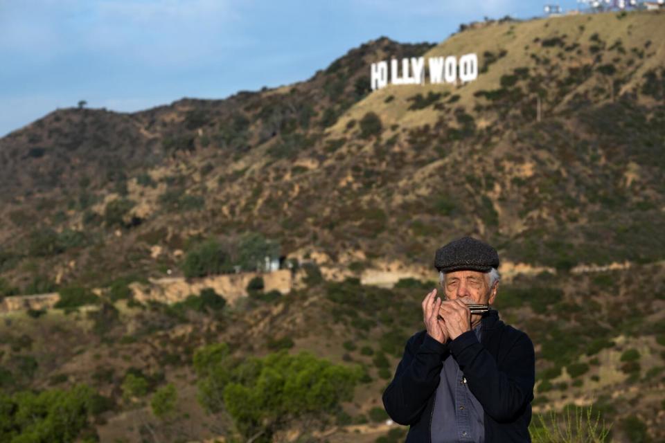 A man plays an harmonica in front of a hill with the "Hollywood" sign.