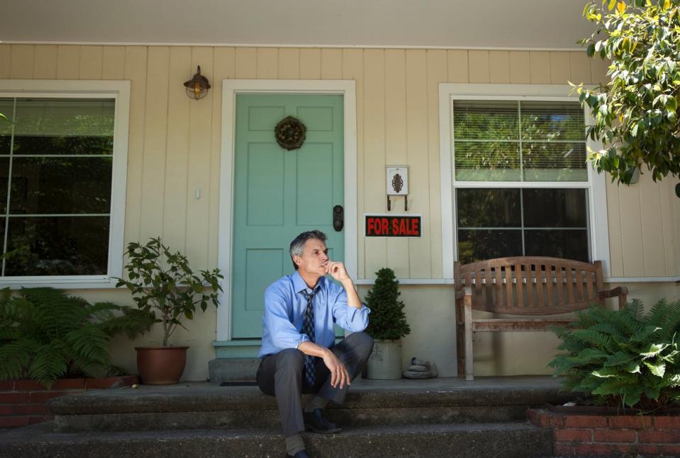 man in suit sits on porch of house foreclosed with for sale sign by door