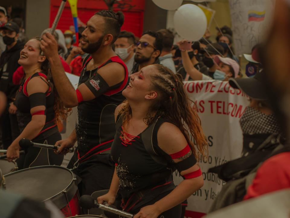 A woman and man dressed in black and red playing drums in a street protest.