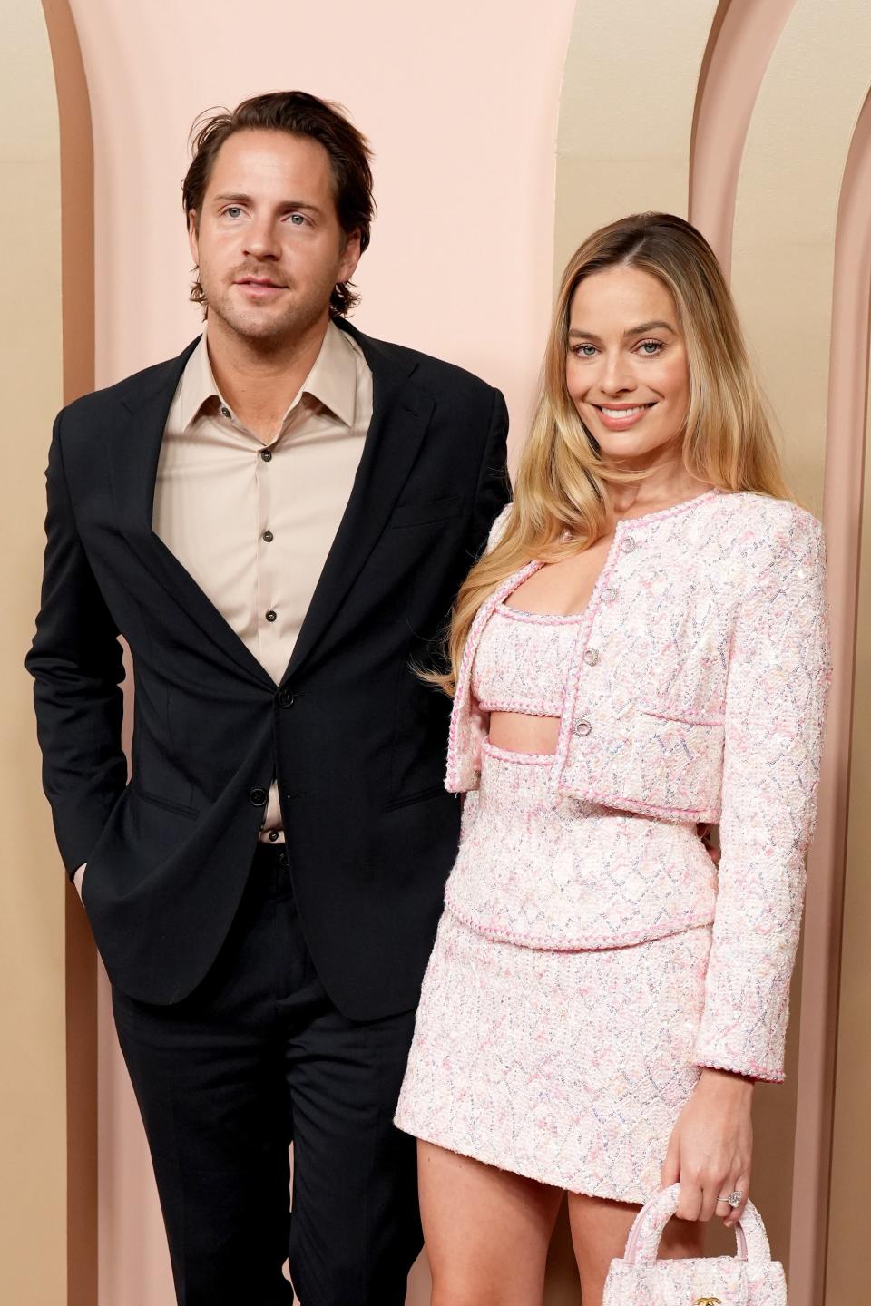 Tom Ackerley and Margot Robbie smile on the red carpet. Margot wears a chic, pink tweed crop top and matching mini skirt with a blazer. Tom wears a black suit