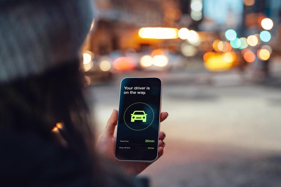 Over the shoulder view of young woman using mobile app on smart phone to arrange taxi service on the street (driving apps to make money) 