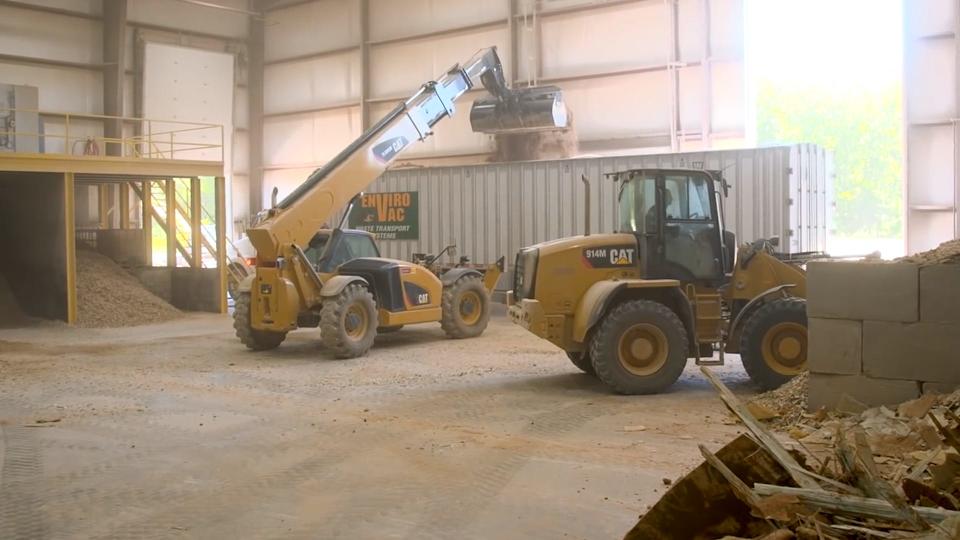 Inside a warehouse, an excavator lowers chunks of wood and fiberglass into a shredding machine.