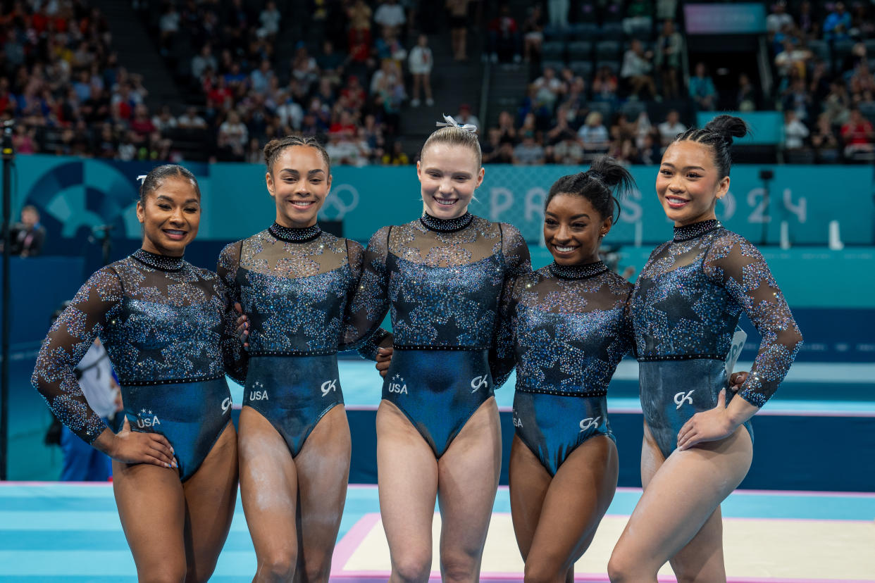  Jordan Chiles, Hezly Rivera, Jade Carey, Simone Biles and Sunisa Lee stand arm in arm in front of fans in stadium seating.