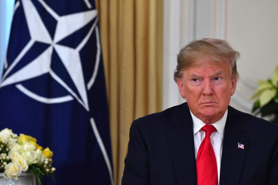Donald Trump glares in a meeting in which the NATO flag sits in the background.
