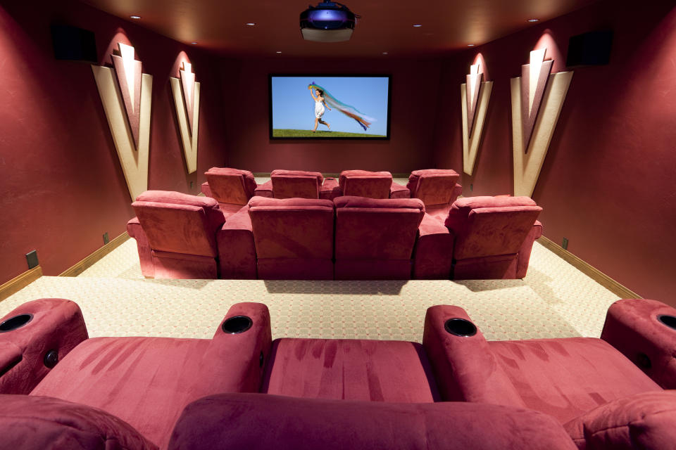 A home theater with red seats arranged in rows facing a large screen displaying an image of a person in a light-colored outfit with a flowing blue cape