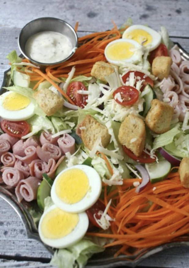 Garden salad with eggs, croutons, vegetables, and ham on a wooden surface with dressing on the side