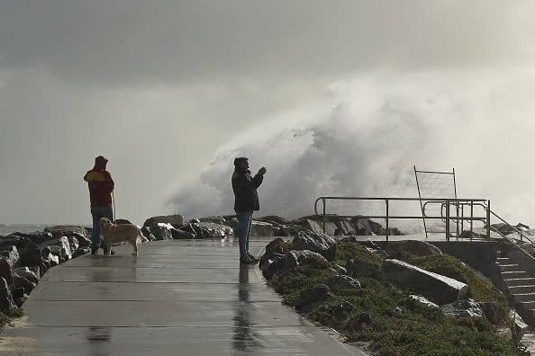 A man takes footage on his phone as a large swell hits the rock groyne at Cottesloe Beach.