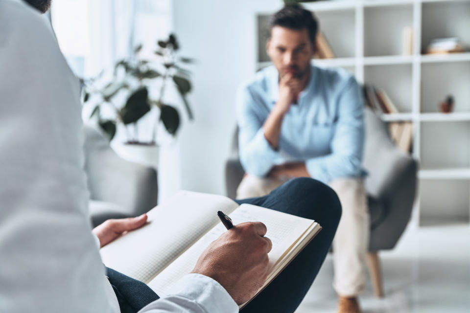 A person attentively listens to another individual who is writing in a notebook during a therapy or consultation session. Shelves and a plant are visible in the background