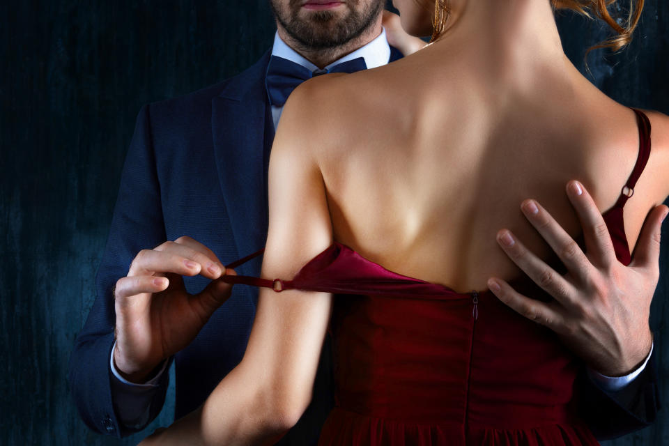 A man in a suit and bow tie adjusts the strap of a woman's red dress on her shoulder