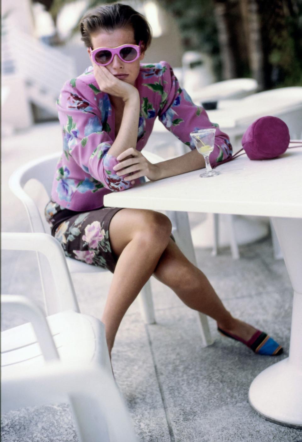 Model Jenny Howarth sitting at a patio table at the Fontainebleau Hilton Resort and Spa in Miami Beach, Florida. She wears a floral printed fitted silk jacket and short skirt, both from Emanuel Ungaro Parallele, pink sunglasses from Christian Lacroix for Optyl Sunglasses, a pink drawstring bag from Prada, and flat color-blocked shoes from Unisa. Hair by Christiaan. Makeup by Sonia Kashuk.