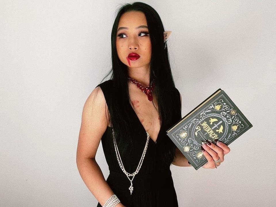 A woman in a black dress with fake blood on her mouth holds a book.