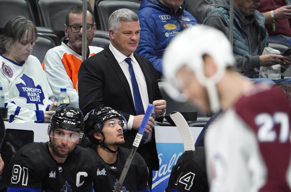 Toronto Maple Leafs head coach Sheldon Keefe, back, looks on from the team box in the second period of an NHL hockey game against the Colorado Avalanche Saturday, Feb. 24, 2024, in Denver. (AP Photo/David Zalubowski)