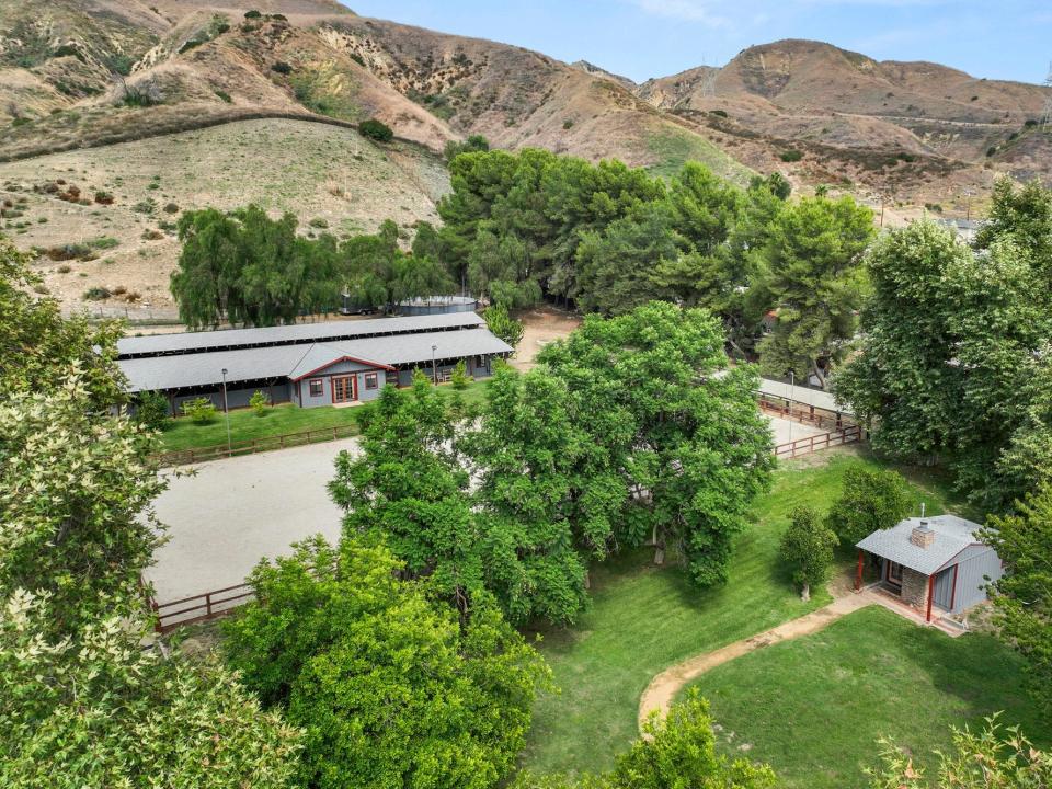 A gray ranch home with large green hills behind it and large trees surrounding it